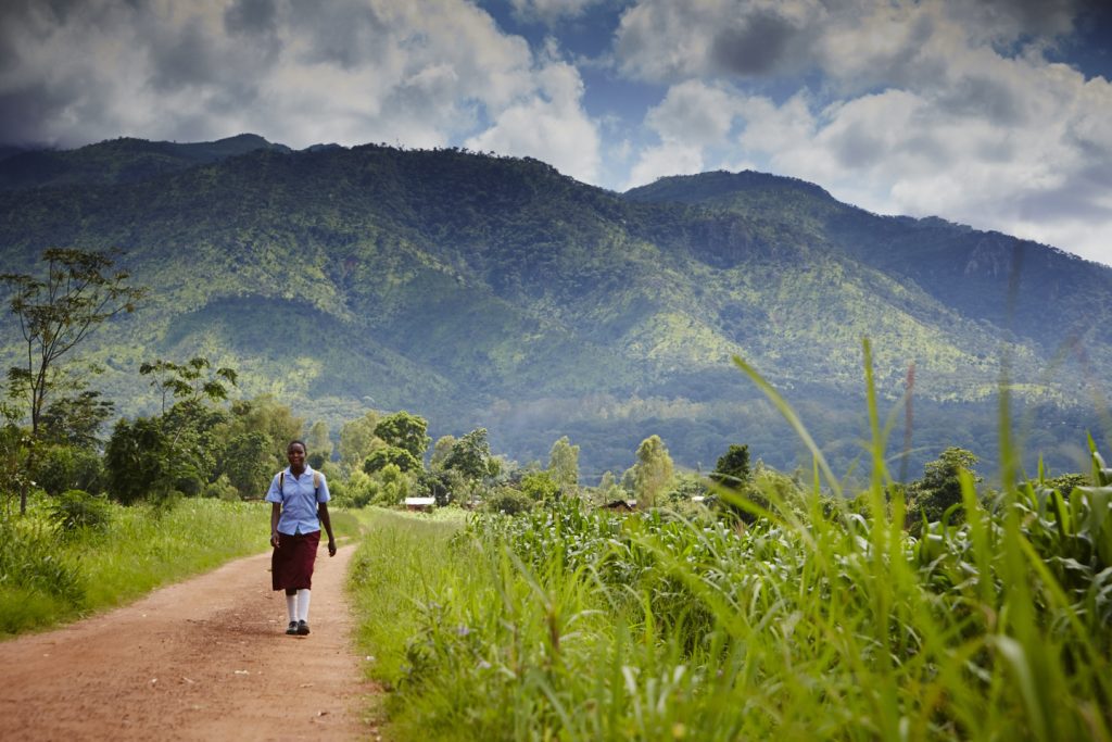 Likwenu, a school girl in uniform, walks along a path with mountains in the background of the image